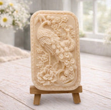 Load image into Gallery viewer, handmade vegan soap featuring a highly detailed relief carving of a peacock with a long, flowing tail, surrounded by delicate cherry blossoms and a large peony, displayed on a wooden easel.