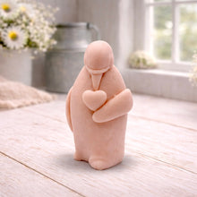 Load image into Gallery viewer, A 3D handmade vegan soap sculpted as a standing penguin tenderly cradling a heart against its chest, displayed on a light wooden surface in a bright farmhouse setting.