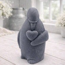 Load image into Gallery viewer, A 3D handmade vegan soap sculpted as a standing penguin tenderly cradling a heart against its chest, displayed on a light wooden surface in a bright farmhouse setting.
