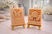 Load image into Gallery viewer, Two rectangular handmade vegan soaps displayed on miniature wooden easels; the left soap features a circular butterfly relief with the words "Embrace Your Dreams," and the right soap features a sunflower relief with the words "Life is Good".