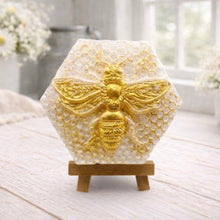 Load image into Gallery viewer, A hexagonal handmade vegan soap featuring a detailed relief of a large queen bee with outstretched wings, highlighted in shimmering gold, set against a textured white honeycomb background and displayed on a miniature wooden easel.