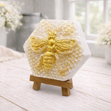 Load image into Gallery viewer, A hexagonal handmade vegan soap featuring a detailed relief of a large queen bee with outstretched wings, highlighted in shimmering gold, set against a textured white honeycomb background and displayed on a miniature wooden easel.