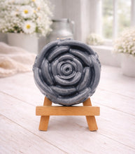 Load image into Gallery viewer, A handmade vegan soap, intricately sculpted in the shape of a large, blooming rose flower with layered petals, displayed on a small wooden easel against a bright win