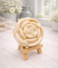 Load image into Gallery viewer, A handmade vegan soap, intricately sculpted in the shape of a large, blooming rose flower with layered petals, displayed on a small wooden easel against a bright win