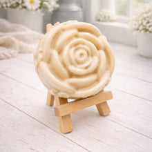 Load image into Gallery viewer, A handmade vegan soap, intricately sculpted in the shape of a large, blooming rose flower with layered petals, displayed on a small wooden easel against a bright win
