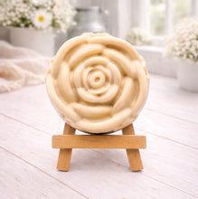 Load image into Gallery viewer, A handmade vegan soap, intricately sculpted in the shape of a large, blooming rose flower with layered petals, displayed on a small wooden easel against a bright win