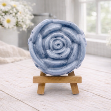 Load image into Gallery viewer, A handmade vegan soap, intricately sculpted in the shape of a large, blooming rose flower with layered petals, displayed on a small wooden easel against a bright win