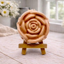 Load image into Gallery viewer, A handmade vegan soap, intricately sculpted in the shape of a large, blooming rose flower with layered petals, displayed on a small wooden easel against a bright win