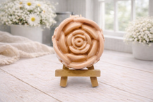 Load image into Gallery viewer, A handmade vegan soap, intricately sculpted in the shape of a large, blooming rose flower with layered petals, displayed on a small wooden easel against a bright win