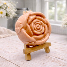 Load image into Gallery viewer, A handmade vegan soap, intricately sculpted in the shape of a large, blooming rose flower with layered petals, displayed on a small wooden easel against a bright win