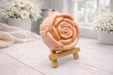 Load image into Gallery viewer, A handmade vegan soap, intricately sculpted in the shape of a large, blooming rose flower with layered petals, displayed on a small wooden easel against a bright win