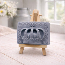 Load image into Gallery viewer, A rectangular handmade vegan soap in a soft tone, featuring a highly detailed relief of a royal crown with floral border accents, displayed on a miniature wooden easel in a bright farmhouse setting.
