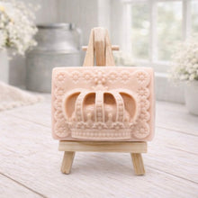 Load image into Gallery viewer, A rectangular handmade vegan soap in a soft tone, featuring a highly detailed relief of a royal crown with floral border accents, displayed on a miniature wooden easel in a bright farmhouse setting.