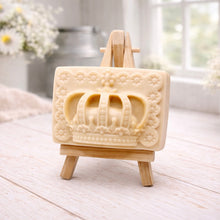 Load image into Gallery viewer, A rectangular handmade vegan soap in a soft tone, featuring a highly detailed relief of a royal crown with floral border accents, displayed on a miniature wooden easel in a bright farmhouse setting.