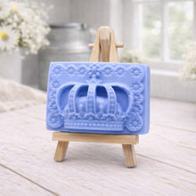 Load image into Gallery viewer, A rectangular handmade vegan soap in a soft tone, featuring a highly detailed relief of a royal crown with floral border accents, displayed on a miniature wooden easel in a bright farmhouse setting.