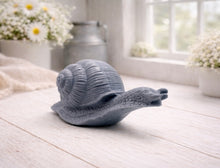 Load image into Gallery viewer, A highly detailed handmade vegan soap in a slate-grey tone, sculpted in the shape of a realistic snail with a prominent spiralled shell and a textured, leathery body, displayed on a light wooden surface.
