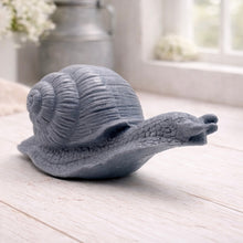Load image into Gallery viewer, A highly detailed handmade vegan soap in a slate-grey tone, sculpted in the shape of a realistic snail with a prominent spiralled shell and a textured, leathery body, displayed on a light wooden surface.