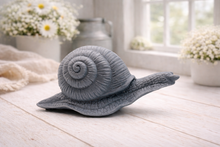 Load image into Gallery viewer, A highly detailed handmade vegan soap in a slate-grey tone, sculpted in the shape of a realistic snail with a prominent spiralled shell and a textured, leathery body, displayed on a light wooden surface.