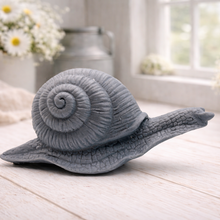 Load image into Gallery viewer, A highly detailed handmade vegan soap in a slate-grey tone, sculpted in the shape of a realistic snail with a prominent spiralled shell and a textured, leathery body, displayed on a light wooden surface.