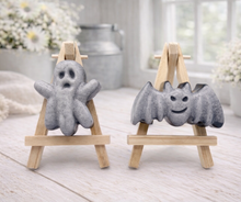 Load image into Gallery viewer, A collection of six handmade vegan Halloween soaps in a mottled grey and black charcoal finish, featuring a smiling bat, three different carved pumpkins (one wearing a witch's hat), a ghost, and a skull, arranged on and around a small wooden easel.