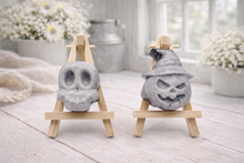 Load image into Gallery viewer, Two vegan soap sculptures of a skull and a jack-o'-lantern on small wooden stands in a bright room.
