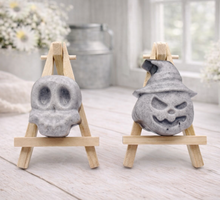 Load image into Gallery viewer, Two vegan soap sculptures of a skull and a jack-o'-lantern on small wooden stands in a bright room.