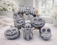 Load image into Gallery viewer, A collection of six handmade vegan Halloween soaps in a mottled grey and black charcoal finish, featuring a smiling bat, three different carved pumpkins (one wearing a witch's hat), a ghost, and a skull, arranged on and around a small wooden easel.
