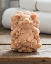 Load image into Gallery viewer, A detailed handmade vegan soap, sculpted as a cheerful pig sitting in a mound of bubbles and holding a scrubbing brush, displayed on a miniature wooden easel against a soft farmhouse window background