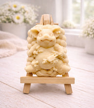 Load image into Gallery viewer, A detailed handmade vegan soap, sculpted as a cheerful pig sitting in a mound of bubbles and holding a scrubbing brush, displayed on a miniature wooden easel against a soft farmhouse window background