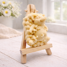 Load image into Gallery viewer, A detailed handmade vegan soap, sculpted as a cheerful pig sitting in a mound of bubbles and holding a scrubbing brush, displayed on a miniature wooden easel against a soft farmhouse window background