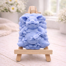 Load image into Gallery viewer, A detailed handmade vegan soap, sculpted as a cheerful pig sitting in a mound of bubbles and holding a scrubbing brush, displayed on a miniature wooden easel against a soft farmhouse window background