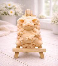 Load image into Gallery viewer, A detailed handmade vegan soap, sculpted as a cheerful pig sitting in a mound of bubbles and holding a scrubbing brush, displayed on a miniature wooden easel against a soft farmhouse window background