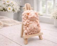 Load image into Gallery viewer, A detailed handmade vegan soap, sculpted as a cheerful pig sitting in a mound of bubbles and holding a scrubbing brush, displayed on a miniature wooden easel against a soft farmhouse window background