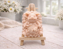 Load image into Gallery viewer, A detailed handmade vegan soap, sculpted as a cheerful pig sitting in a mound of bubbles and holding a scrubbing brush, displayed on a miniature wooden easel against a soft farmhouse window background