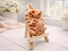 Load image into Gallery viewer, A detailed handmade vegan soap, sculpted as a cheerful pig sitting in a mound of bubbles and holding a scrubbing brush, displayed on a miniature wooden easel against a soft farmhouse window background