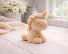 Load image into Gallery viewer, A 3D handmade vegan soap sculpted into a cute sitting unicorn with a spiralled horn, a smiling expression with a closed winking eye, and a bubbly mane and tail, displayed on a light wooden surface in a bright farmhouse setting.