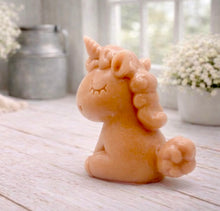 Load image into Gallery viewer, A 3D handmade vegan soap sculpted into a cute sitting unicorn with a spiralled horn, a smiling expression with a closed winking eye, and a bubbly mane and tail, displayed on a light wooden surface in a bright farmhouse setting.