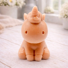 Load image into Gallery viewer, A 3D handmade vegan soap sculpted into a cute sitting unicorn with a spiralled horn, a smiling expression with a closed winking eye, and a bubbly mane and tail, displayed on a light wooden surface in a bright farmhouse setting.