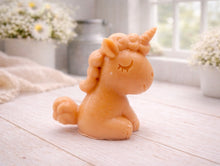 Load image into Gallery viewer, A 3D handmade vegan soap sculpted into a cute sitting unicorn with a spiralled horn, a smiling expression with a closed winking eye, and a bubbly mane and tail, displayed on a light wooden surface in a bright farmhouse setting.