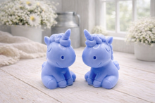 Load image into Gallery viewer, A 3D handmade vegan soap sculpted into a cute sitting unicorn with a spiralled horn, a smiling expression with a closed winking eye, and a bubbly mane and tail, displayed on a light wooden surface in a bright farmhouse setting.