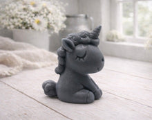 Load image into Gallery viewer, A 3D handmade vegan soap sculpted into a cute sitting unicorn with a spiralled horn, a smiling expression with a closed winking eye, and a bubbly mane and tail, displayed on a light wooden surface in a bright farmhouse setting.
