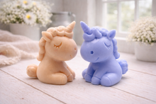 Load image into Gallery viewer, A 3D handmade vegan soap sculpted into a cute sitting unicorn with a spiralled horn, a smiling expression with a closed winking eye, and a bubbly mane and tail, displayed on a light wooden surface in a bright farmhouse setting.