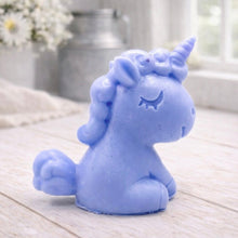 Load image into Gallery viewer, A 3D handmade vegan soap sculpted into a cute sitting unicorn with a spiralled horn, a smiling expression with a closed winking eye, and a bubbly mane and tail, displayed on a light wooden surface in a bright farmhouse setting.