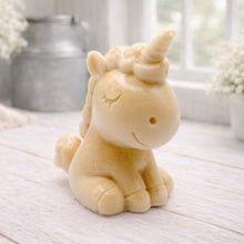 Load image into Gallery viewer, A 3D handmade vegan soap sculpted into a cute sitting unicorn with a spiralled horn, a smiling expression with a closed winking eye, and a bubbly mane and tail, displayed on a light wooden surface in a bright farmhouse setting.