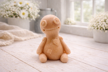 Load image into Gallery viewer, A handmade vegan soap gift in the shape of a stitched voodoo doll with button eyes and a small patch on its chest, sitting on a light wooden surface.