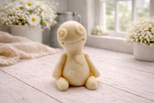 Load image into Gallery viewer, A handmade vegan soap gift in the shape of a stitched voodoo doll with button eyes and a small patch on its chest, sitting on a light wooden surface.