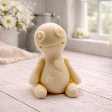 Load image into Gallery viewer, A handmade vegan soap gift in the shape of a stitched voodoo doll with button eyes and a small patch on its chest, sitting on a light wooden surface.