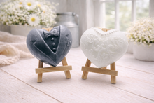 Load image into Gallery viewer, A pair of handmade vegan soaps shaped like hearts and displayed on miniature wooden easels; one heart is textured like a grey tuxedo with a bow tie and buttons, and the other is a white bridal gown heart with intricate lace patterns and a pearl necklace detail.