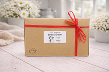 Load image into Gallery viewer, Gift box with a red ribbon labeled 'Bride & Groom' on a wooden surface with flowers in the background.