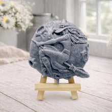 Load image into Gallery viewer, A circular handmade vegan soap featuring a detailed high-relief carving of a witch wearing a pointed hat, flying on a broomstick across a cratered moon, displayed on a miniature wooden easel.